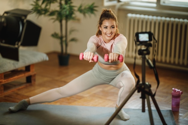 An attractive young woman doing exercises with weight at her home and using camera to record her vlog about healthy living.