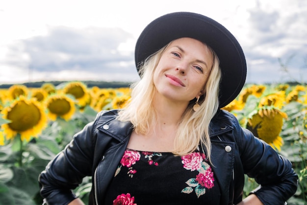 Attractive young woman in dress black leather jacket and hat is posing in field of sunflowers