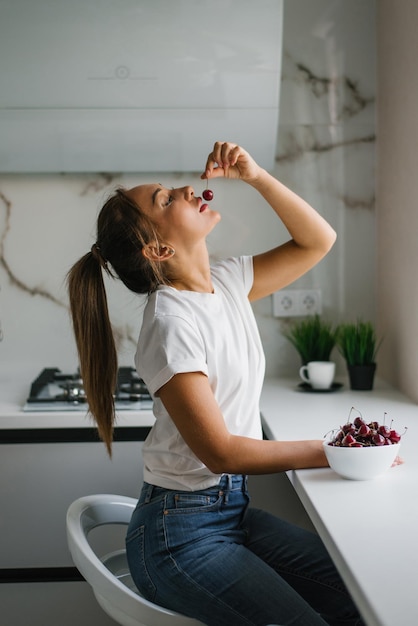 Attraente giovane donna mangia ciliegie fresche bacche sane seduto a casa in cucina