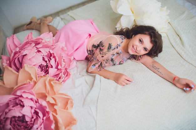 Attractive young woman in elegant dress sitting in interior decorated with flowers. Beautiful fresh smiling girl posing with flowers