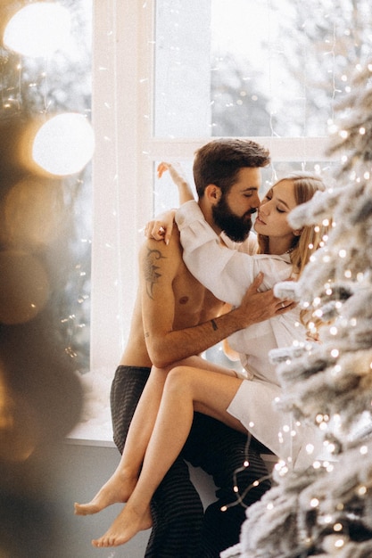 自宅でクリスマスツリーの近くに上半身裸のボーイフレンドを抱きしめる魅力的な若い女性