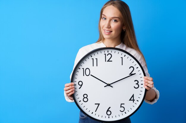 Attractive young woman holding big wall clock against blue background