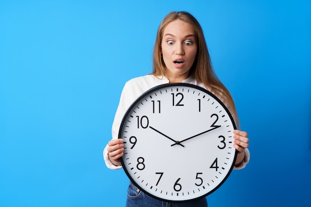 Attractive young woman holding big wall clock against blue background