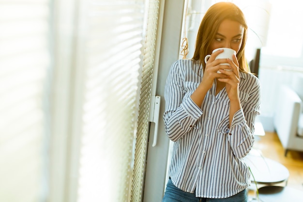 Photo attractive young woman holding cup with hot tea or coffee and looking at the sunrise standing near the window in room