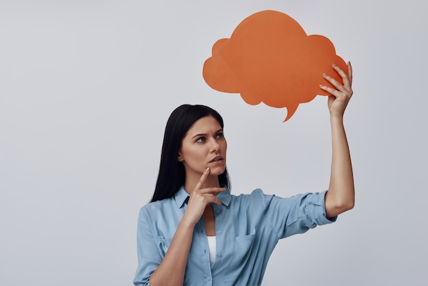 Attractive young woman holding speech bubble while standing against grey wall