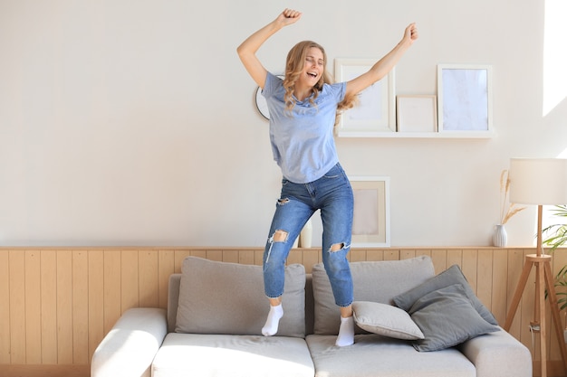 Attractive young woman is having fun at home. Girl is jumping and dancing on sofa.