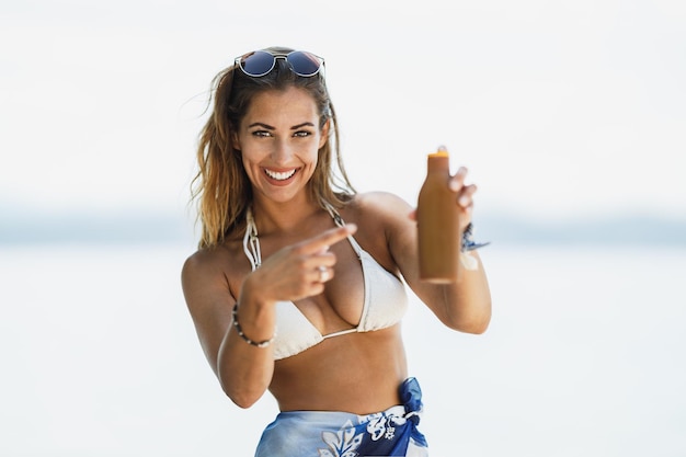 An attractive young woman is holding sun protecion cream and enjoying time on the beach. Selective focus. Looking at camera.