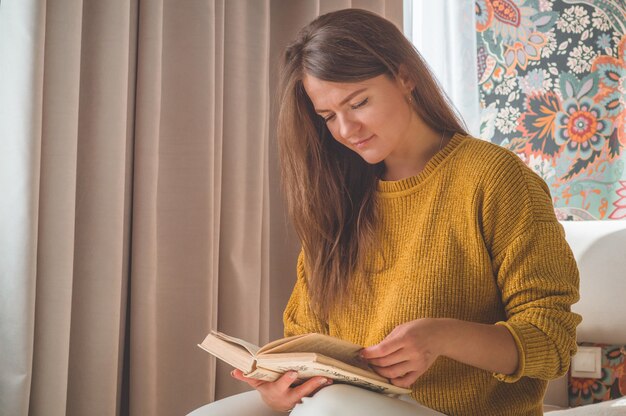 La giovane donna attraente sta leggendo un libro a casa