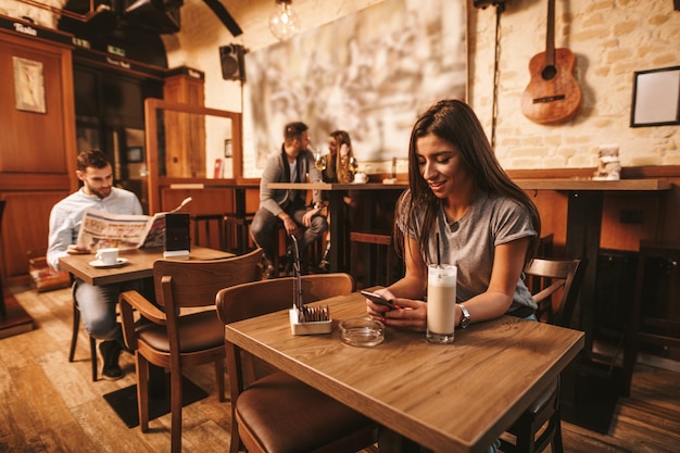 Attraente giovane donna sta leggendo buone notizie sullo smartphone mentre è seduto al bar.