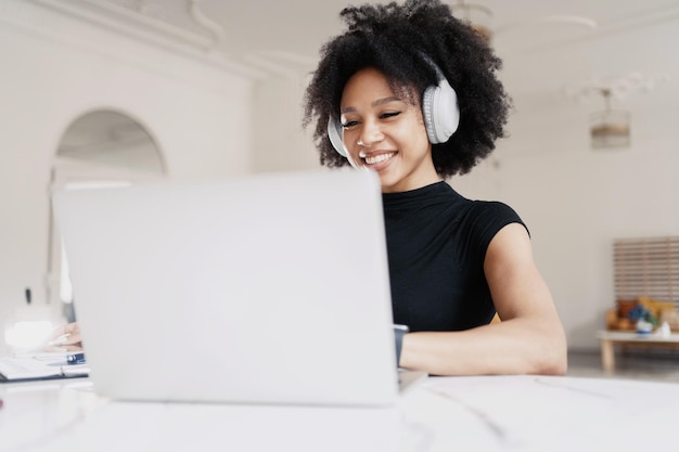 An attractive young woman is smiling sitting at her workplace and using her laptop and headphones