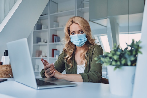 Attractive young woman in protective face mask sitting at the desk in office