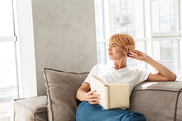 Привлекательная молодая женщина, читающая книгу, сидя на диване у себя дома