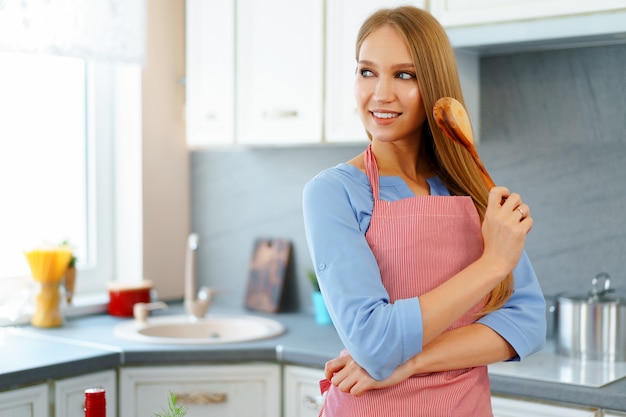 Attraente giovane donna in grembiule rosso in piedi nella sua cucina, ritratto di donna