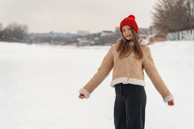 赤いニット帽とベージュのシープスキン コートを着た魅力的な若い女性冬の雪に覆われた公園を歩く