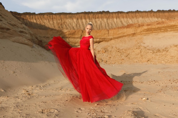 Attractive young woman in red waving dress with flying fabric on the background of sands career.