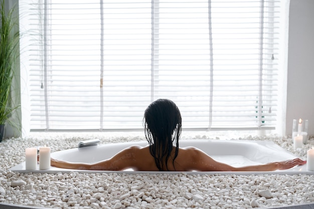 Attractive young woman relax in a bubble bath, back view. Female person in bathtub, beauty and health care in spa, wellness treathment in bathroom, pebbles and candles on background