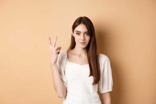 Attractive young woman show fingers number two, smiling and looking confident, standing on beige background.