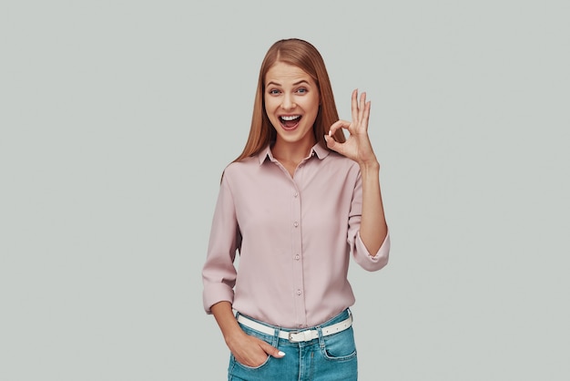 Attractive young woman showing OK symbol and smiling while standing against grey background