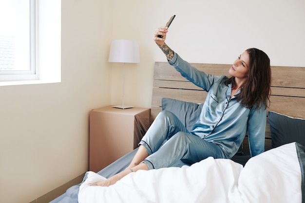 Attractive young woman in silk pajamas sitting on bed and taking selfie after waking up