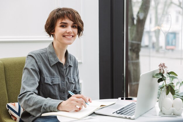 Привлекательная молодая женщина сидит за столом в кафе в помещении, работает на портативном компьютере, анализирует документы