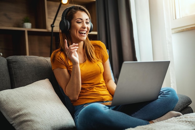 Attraente giovane donna seduta a gambe incrociate sul divano e usando il suo laptop e le cuffie per fare una videochiamata con qualcuno a casa.