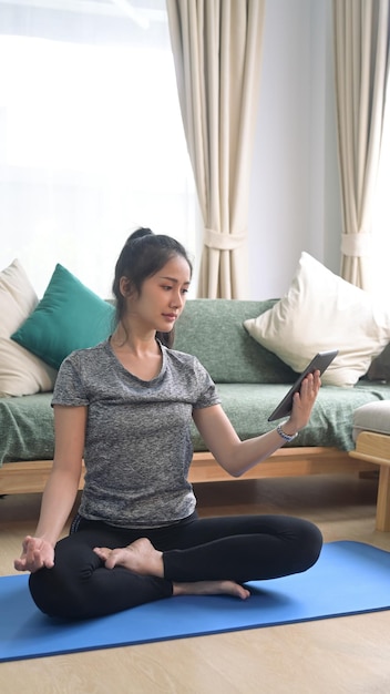 Attractive young woman sitting on yoga mat and using smart phone for watching online workout training class video at home