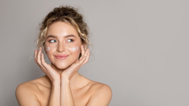 Attractive young woman touching her smooth cheeks applying moisturizer cream looking aside at free