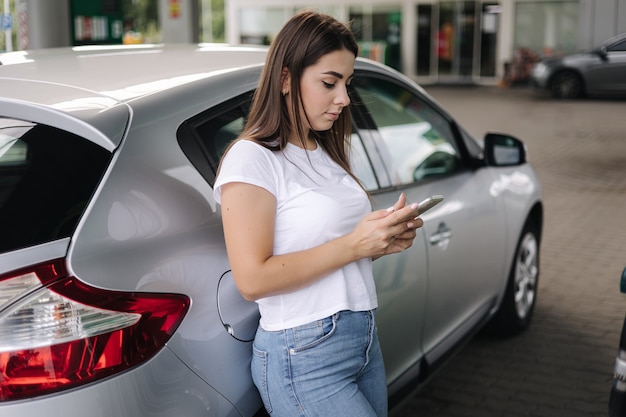 彼女の電話を使用して、ガスを待っているガソリンスタンドの女性で彼女の車に寄りかかった魅力的な若い女性