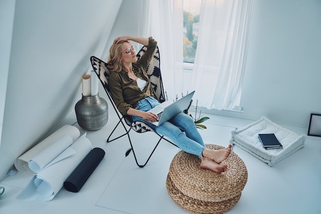 Привлекательная молодая женщина, используя ноутбук, сидя в удобном кресле у себя дома