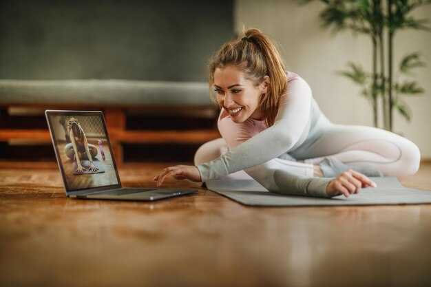 An attractive young woman using laptop while stretching after workout at home during the day.