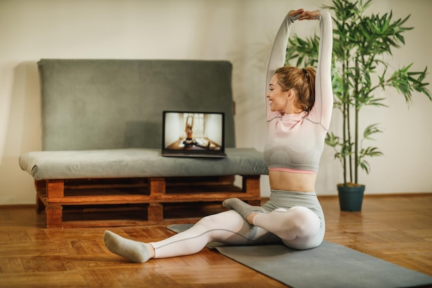 An attractive young woman using laptop while stretching after workout at home.