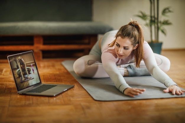 집에서 운동 후 스트레칭을 하는 동안 노트북을 사용하는 매력적인 젊은 여성.