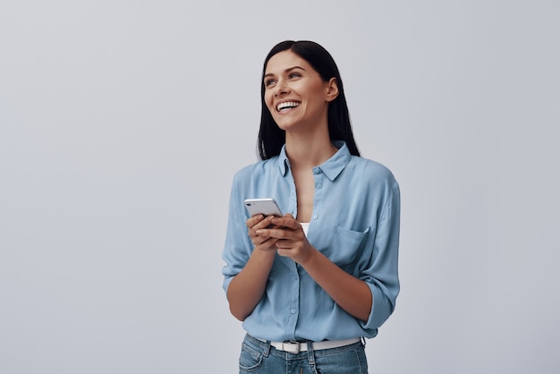 Photo attractive young woman using smart phone and smiling while standing against grey wall