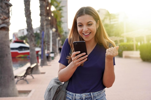 日没時に屋外で良いニュースを祝う彼女のスマートフォンを見ている魅力的な若い女性