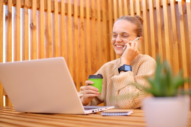 屋外のカフェに座ってラップトップでコーヒーを飲みながら携帯電話で話し、フリーランスの仕事を楽しんでいるベージュのセーターを着た魅力的な若い女性