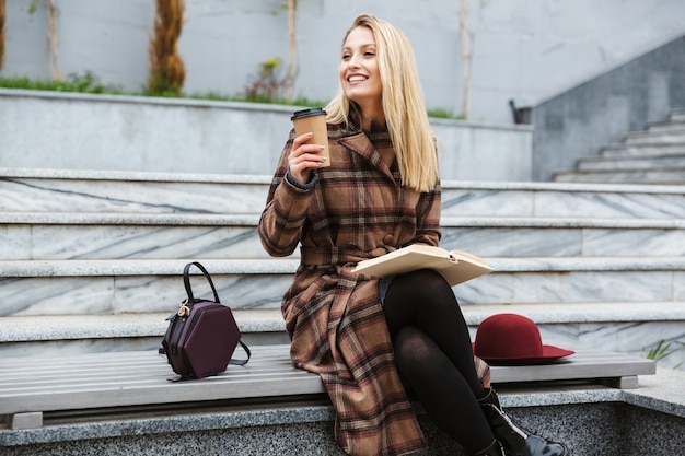 Attractive young woman wearing coat sitting outdoors, reading a book, holding takeaway cup