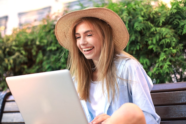 도시 거리의 벤치에 앉아 노트북 작업을 하는 여름 옷을 입은 매력적인 젊은 여성.