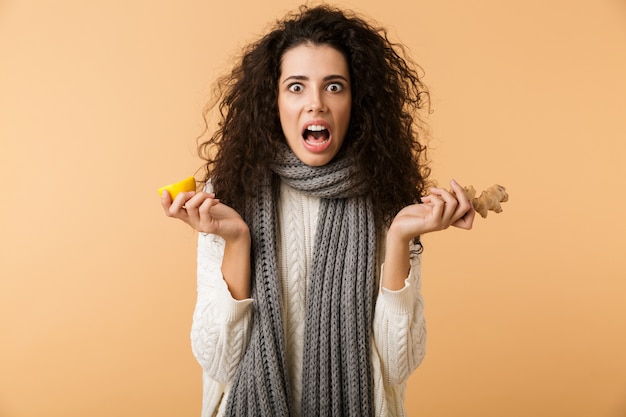 Attractive young woman wearing winter scarf showing ginger and lemon while standing isolated over white wall