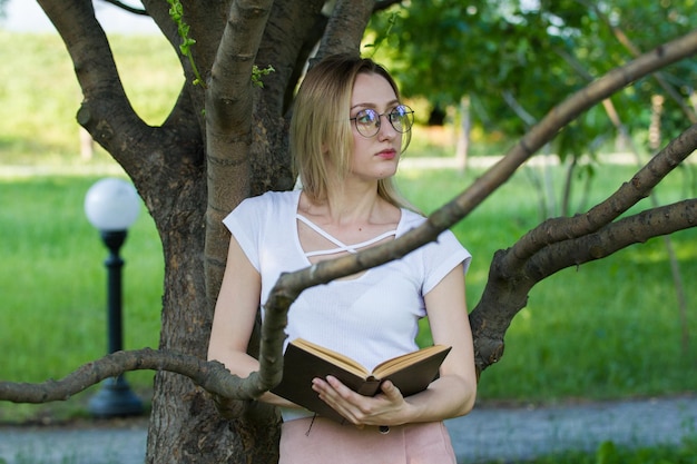 Attractive young woman with a book in the hands in the park leaning on a tree branch, horizontal