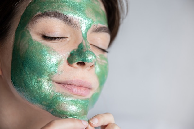 Attraente giovane donna con una maschera cosmetica verde sul viso e in una veste bianca su sfondo grigio, il concetto di trattamenti termali a casa, copia spazio.