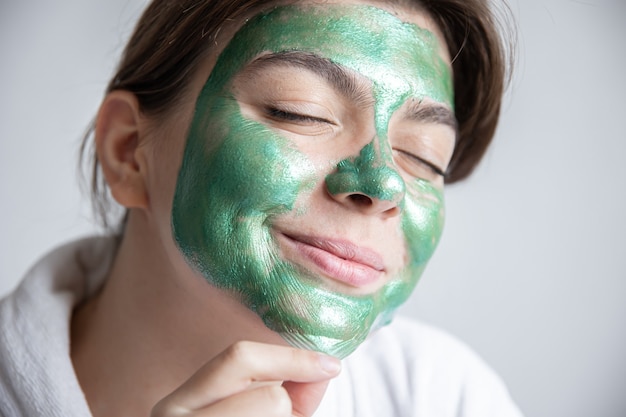 Attractive young woman with a green cosmetic mask on her face and in a white robe on a gray background, the concept of spa treatments at home.