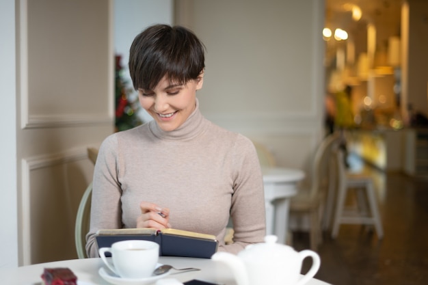 Attractive young woman with pen in hand thinking on plans and writing list to do in notepad enjoying recreation time in stylish cafe