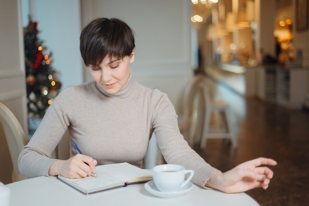 Attractive young woman with pen in hand thinking on plans and writing list to do in notepad enjoying recreation time in stylish cafe