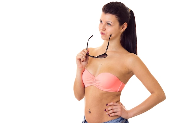 Attractive young woman with ponytail in pink bra and denim shorts with black sunglasses on white background in studio