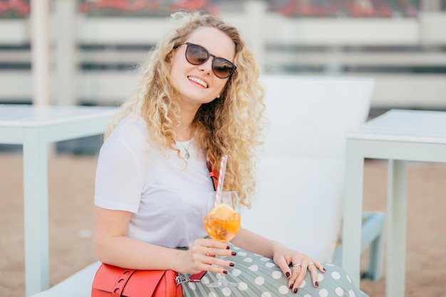 Attractive young woman with wavy hair wears sunglasses has good rest during summer time holds fresh beverage has friendly smile looks positively at camera People drink recreation concept