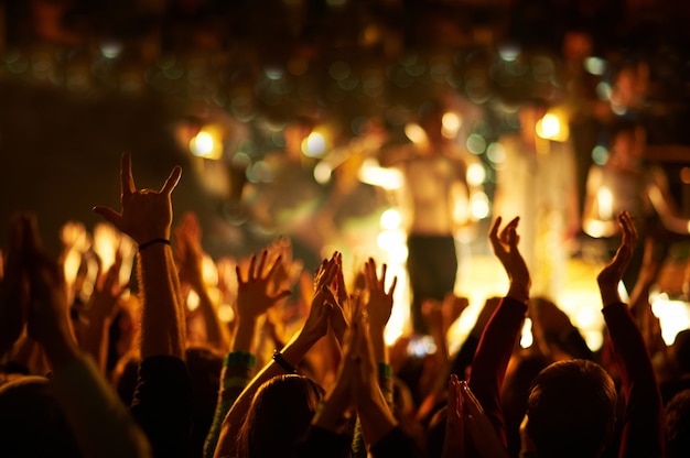 Audience with hands raised at a music festival and lights streaming down from above the stage.