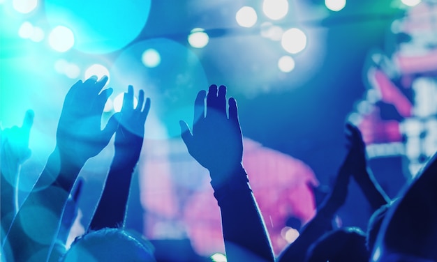 Audience with hands raised at a music festival and lights streaming down from above the stage.