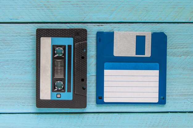 Photo an audio cassette in a black plastic case and a blue floppy disk lie on a blue wooden background