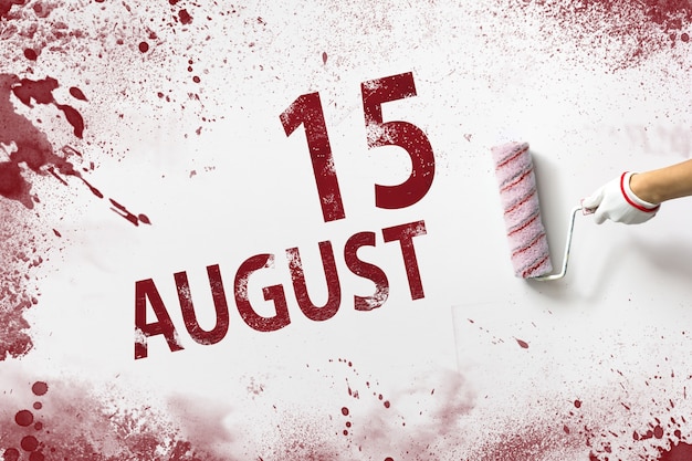 August 15th. day 15 of month, calendar date. the hand holds a\
roller with red paint and writes a calendar date on a white\
background. summer month, day of the year concept.