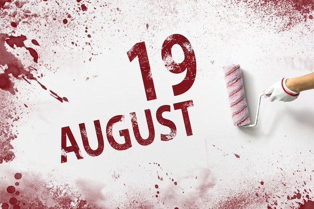 August 19th. day 19 of month, calendar date. the hand holds a\
roller with red paint and writes a calendar date on a white\
background. summer month, day of the year concept.